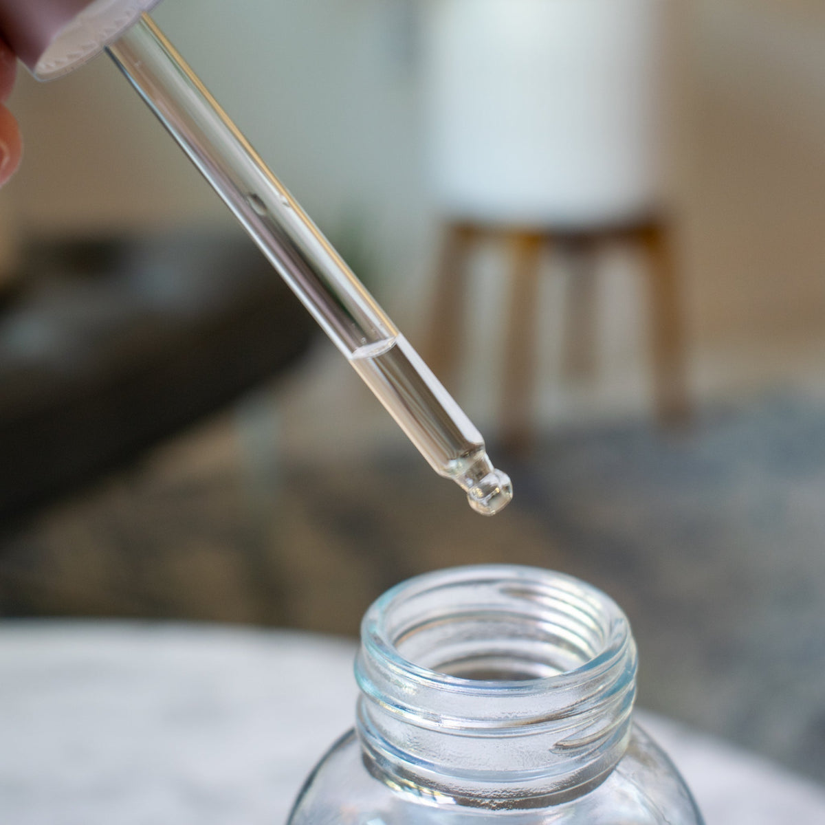 Clear glass rounded tipped dropper above a glass bottle on a blurred background.